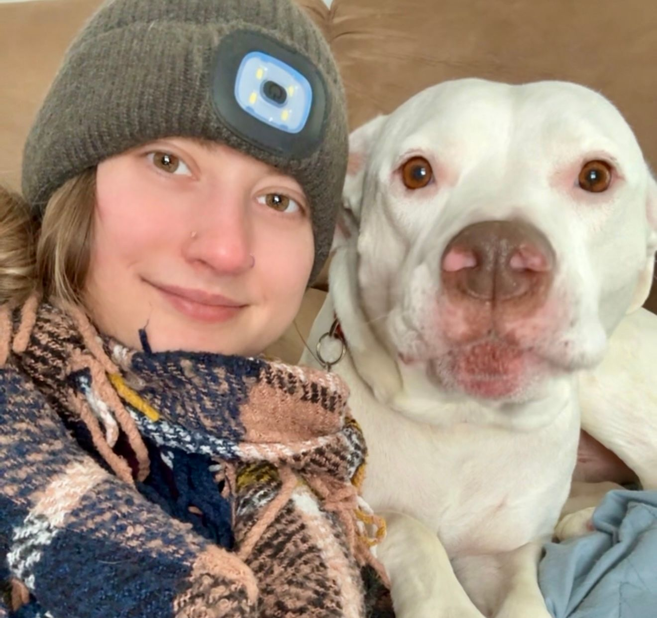 Lex with a beanie and scarf cuddles a white dog, both looking at the camera.