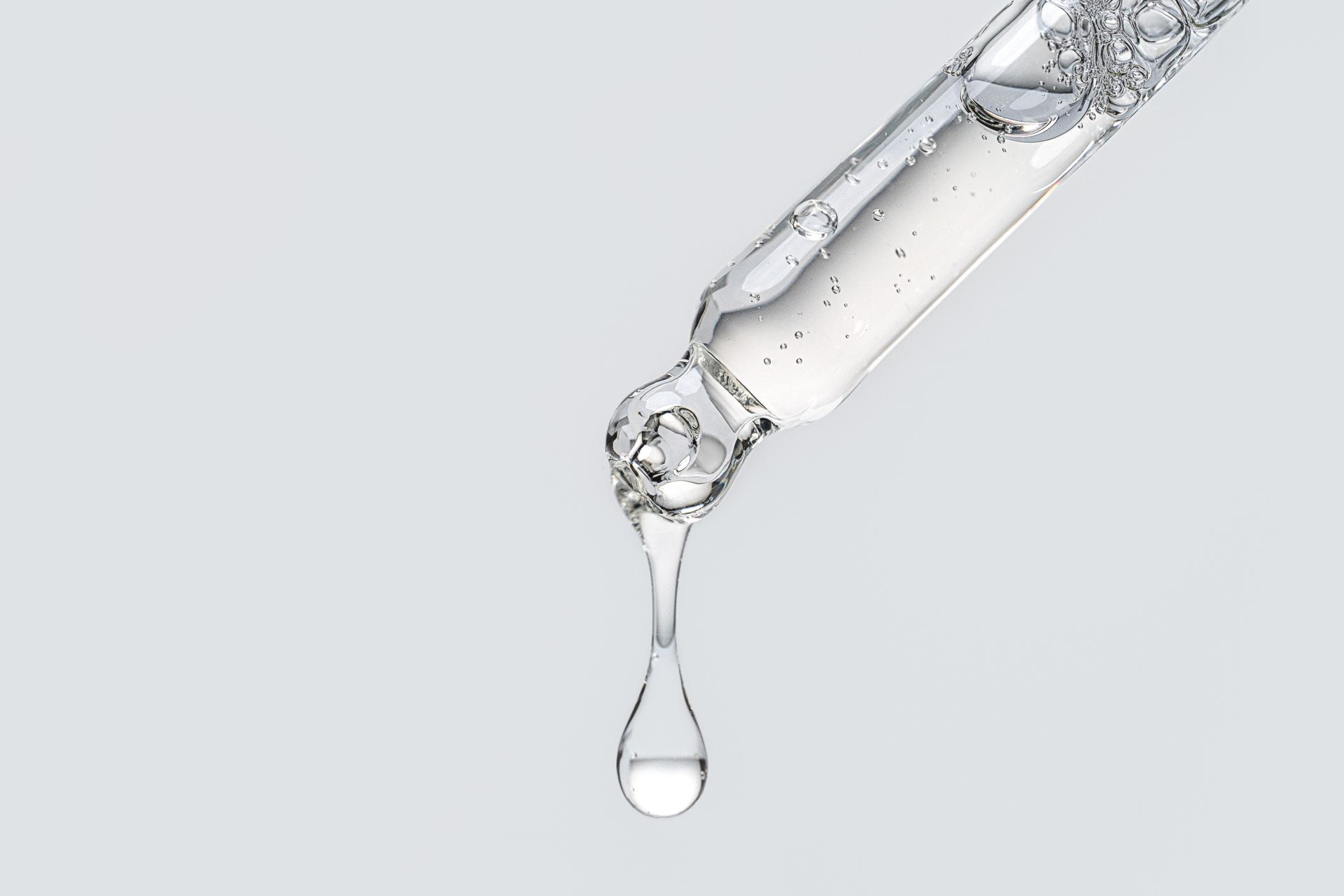 Dropper with clear liquid, a single drop about to fall. Light background.
