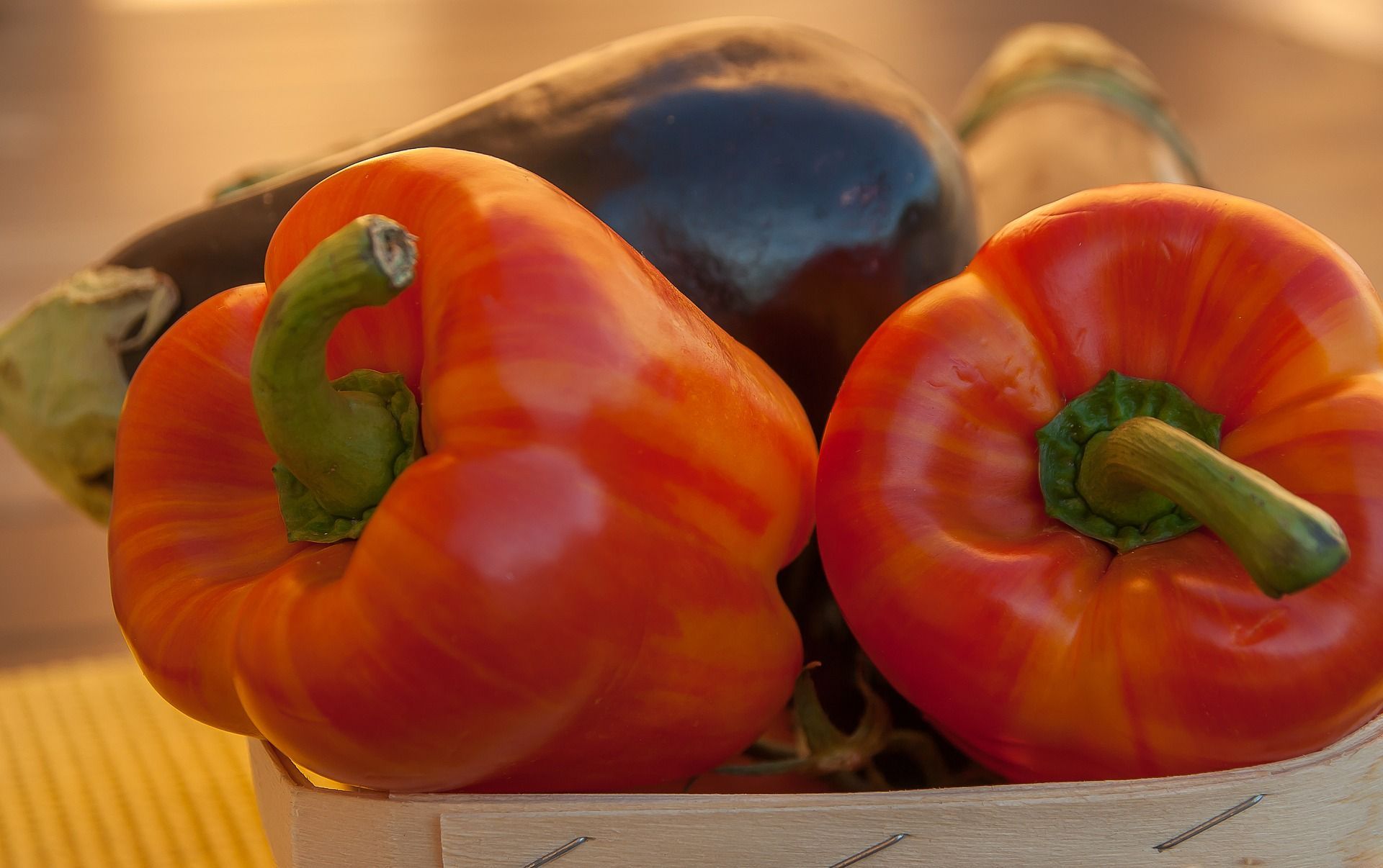 Two red bell peppers in a wooden basket, with a dark eggplant in the background.