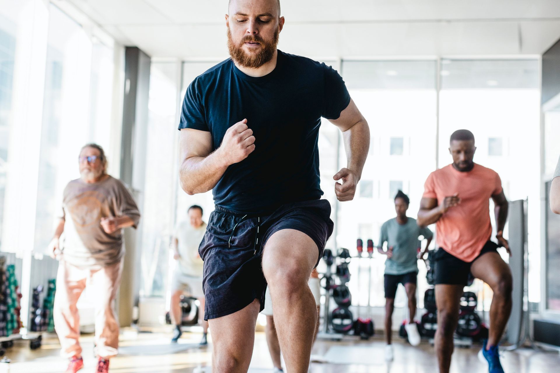 Group of people doing cardio in a gym, led by a bald man in black shirt and shorts.