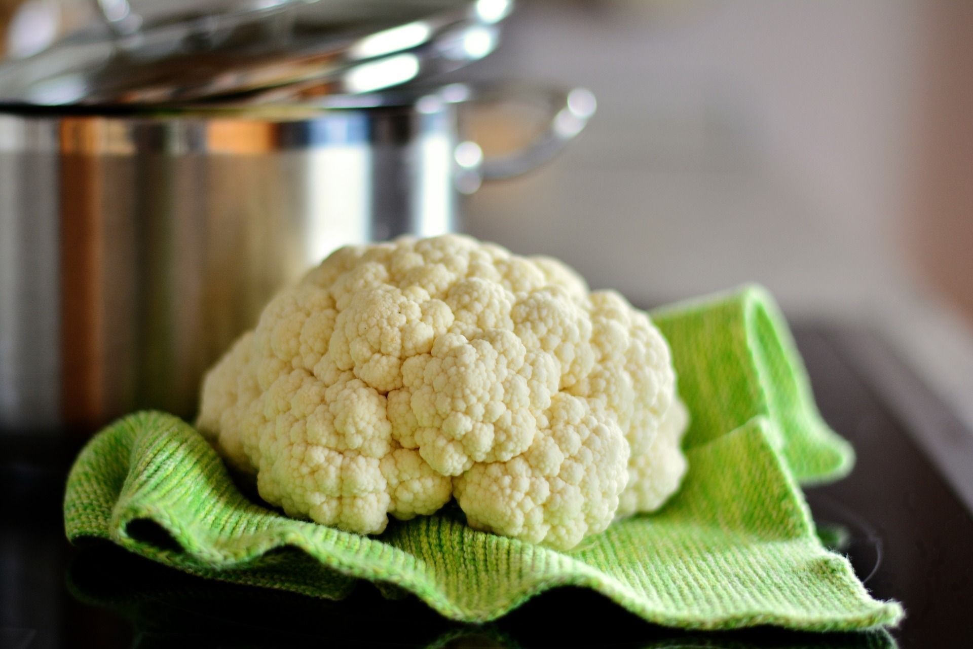 Cauliflower head on green cloth next to a stainless steel pot, indoor kitchen setting.