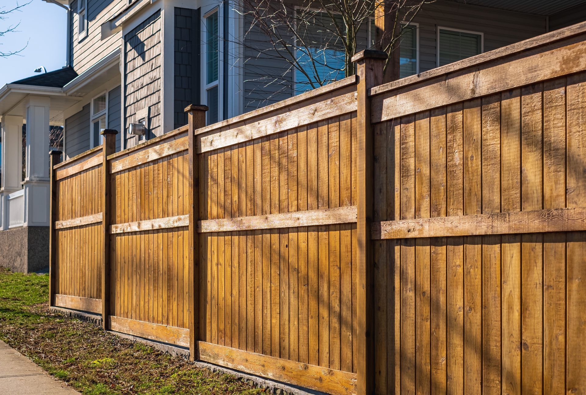 A tall wooden fence with vertical planks and horizontal support beams runs along the side of a house on a sunny day.