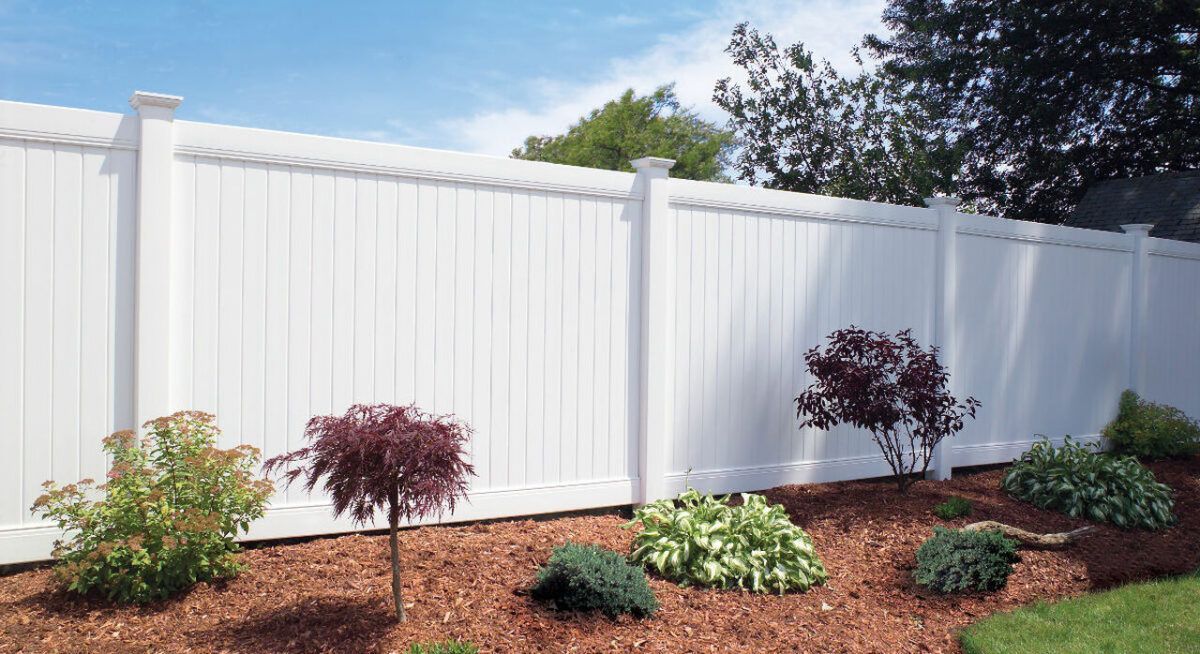 A white vinyl privacy fence with lattice-style trim runs along a garden bed filled with shrubs and mulch under a blue sky.