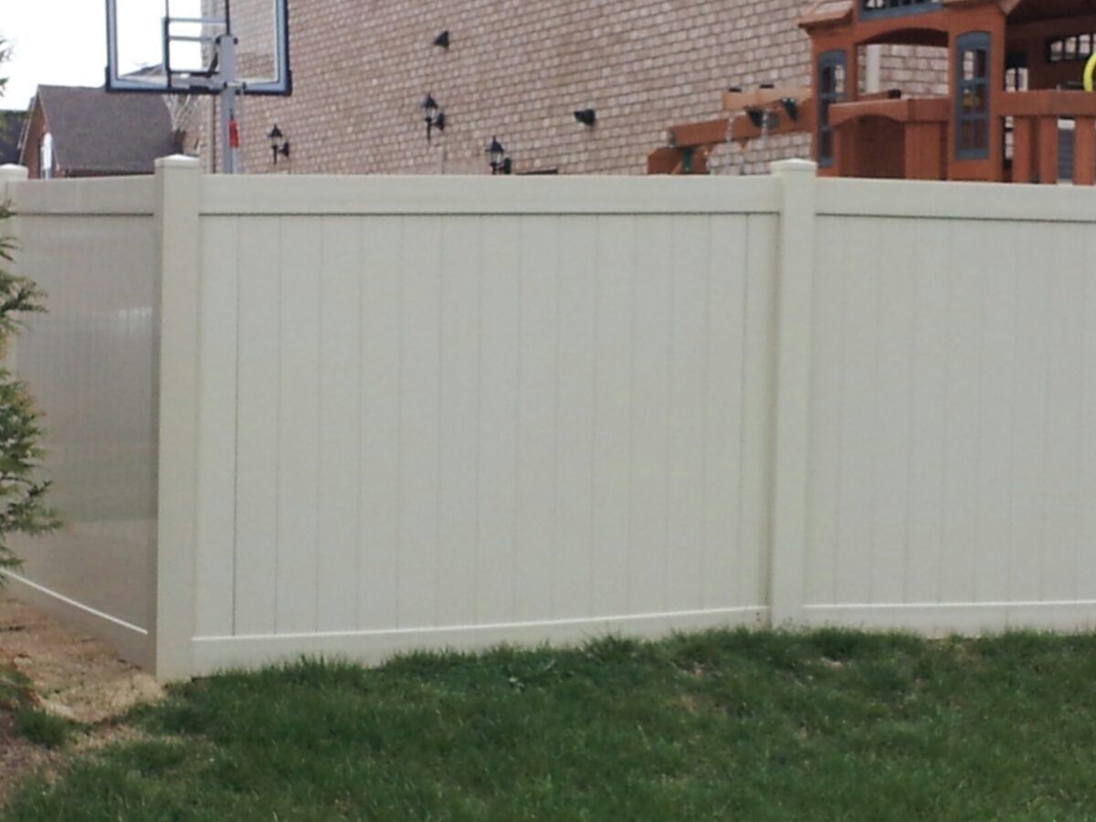Off-white vinyl privacy fence panels installed in a backyard with a brick house, basketball hoop, and playset behind it.