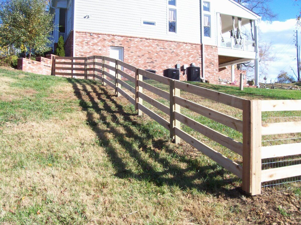 A wooden post-and-rail fence runs along a sloping grassy lawn in front of a brick and white house.