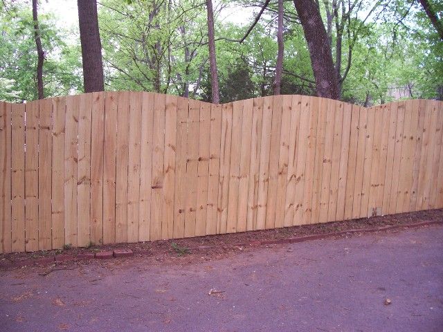 A wood privacy fence with a scalloped top edge, set against a background of trees.
