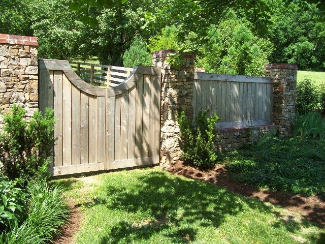 A wooden gate with a curved cutout set into a stone wall, surrounded by lush green foliage and trees in a garden setting.