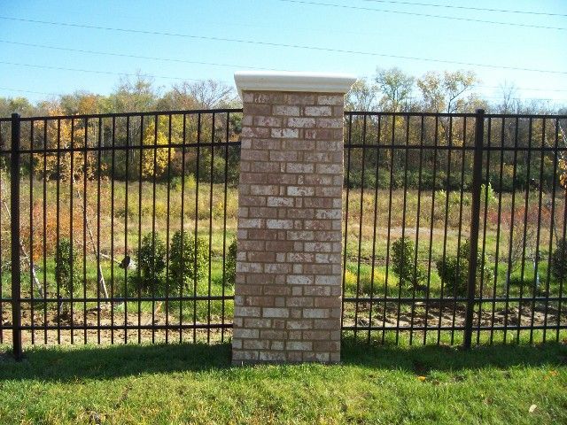 A tall brick pillar stands in the center of a black metal fence, with a background of trees and grass.
