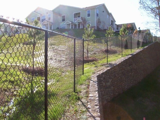 A black chain-link fence runs along a stone border near a grassy hill, with a large residential building in the background.
