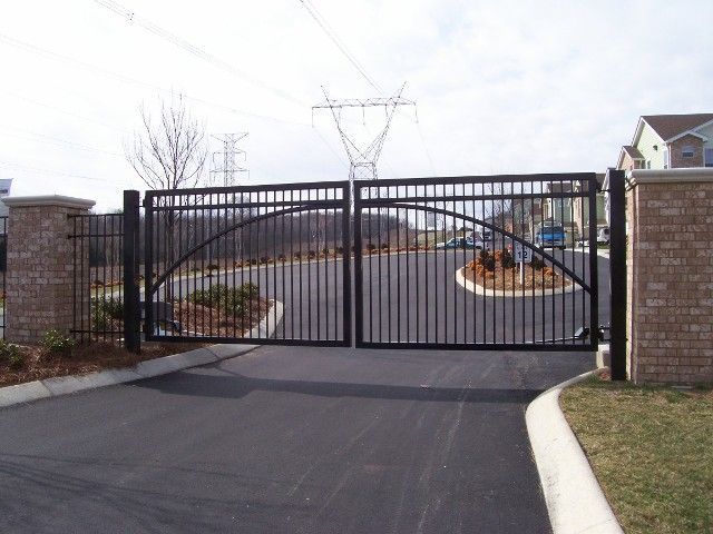 A black metal dual-swing gate with an arched design sits between two brick pillars at a paved residential entrance.