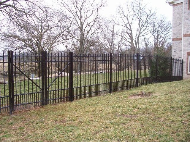 A black metal fence runs across a lawn beside a brick house with bare trees in the background.