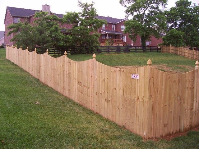 A new wooden scalloped-top privacy fence installed on a residential lawn with houses in the background.