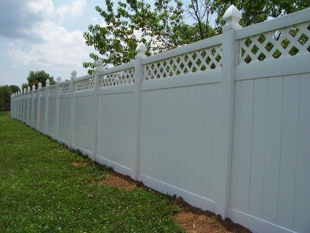 A long, white vinyl privacy fence with a decorative lattice top, stretching across a grassy lawn under a blue sky.