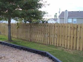 A wooden picket fence stretches across a green lawn in front of a house, with a tree and a black landscape border nearby.
