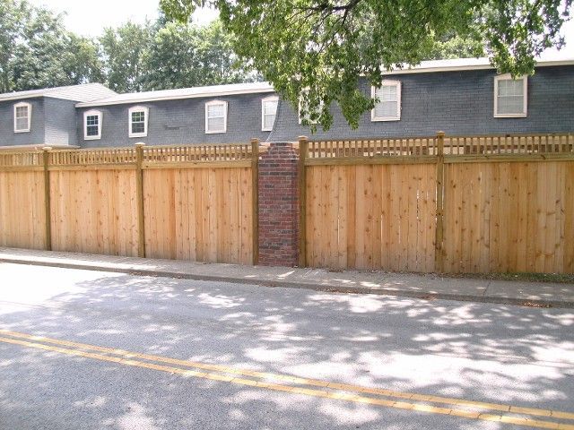 A wooden privacy fence with a decorative brick column section in front of a gray multi-unit building and green trees.