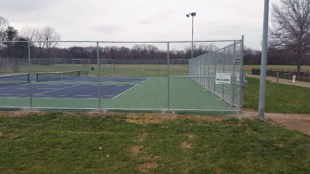A chain-link fence surrounds a tennis court with a blue playing surface and green surrounding area under a cloudy sky.