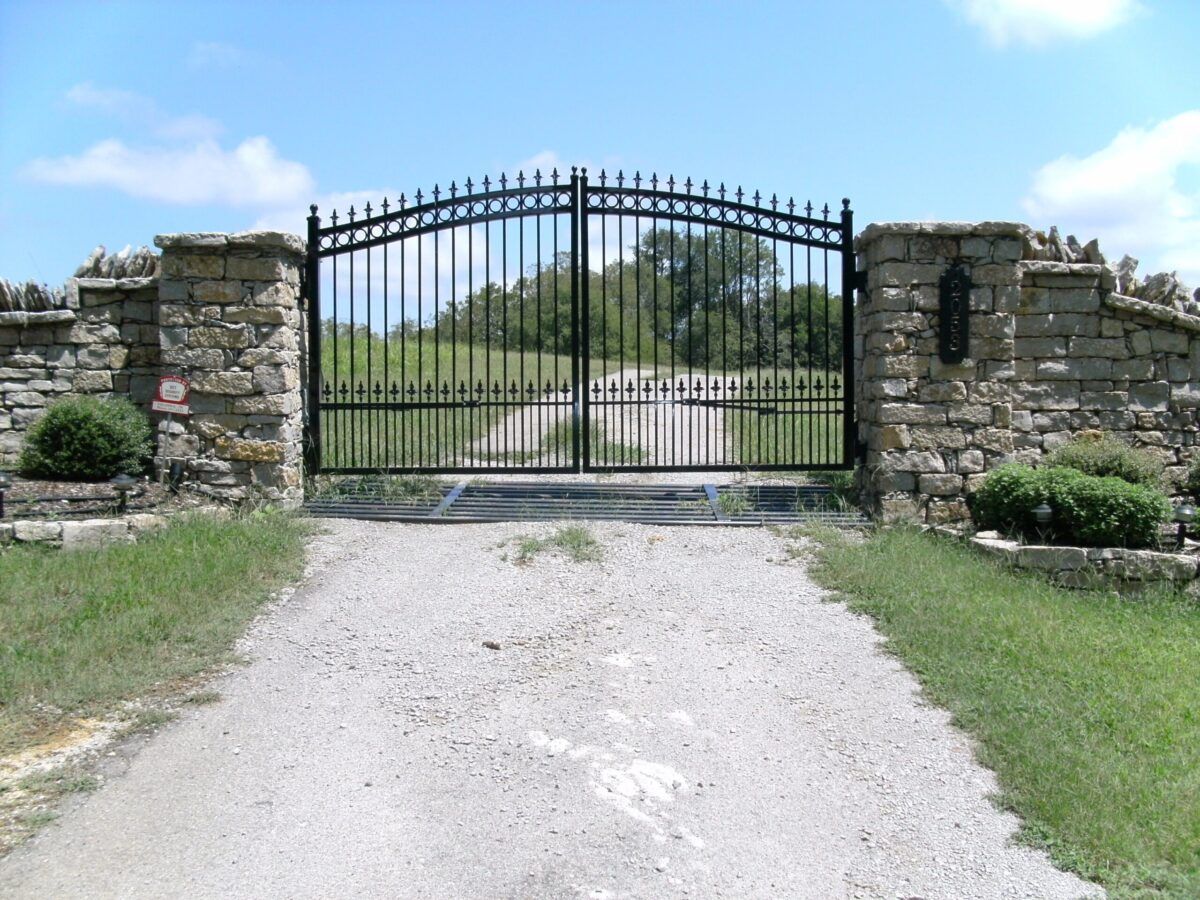 A black metal driveway gate set between stone pillars at the entrance of a gravel path.
