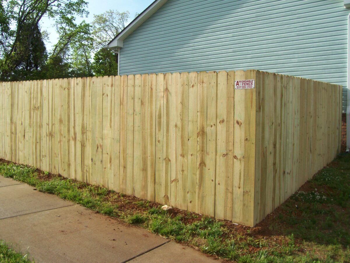 A light-colored wooden privacy fence stands at the corner of a house next to a concrete sidewalk.