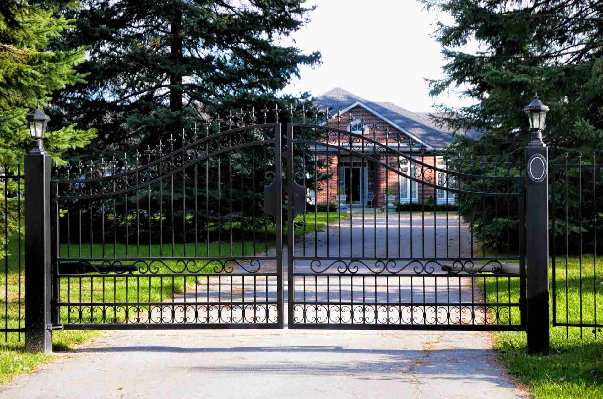 A black iron gate stands across a driveway in front of a brick house surrounded by pine trees.