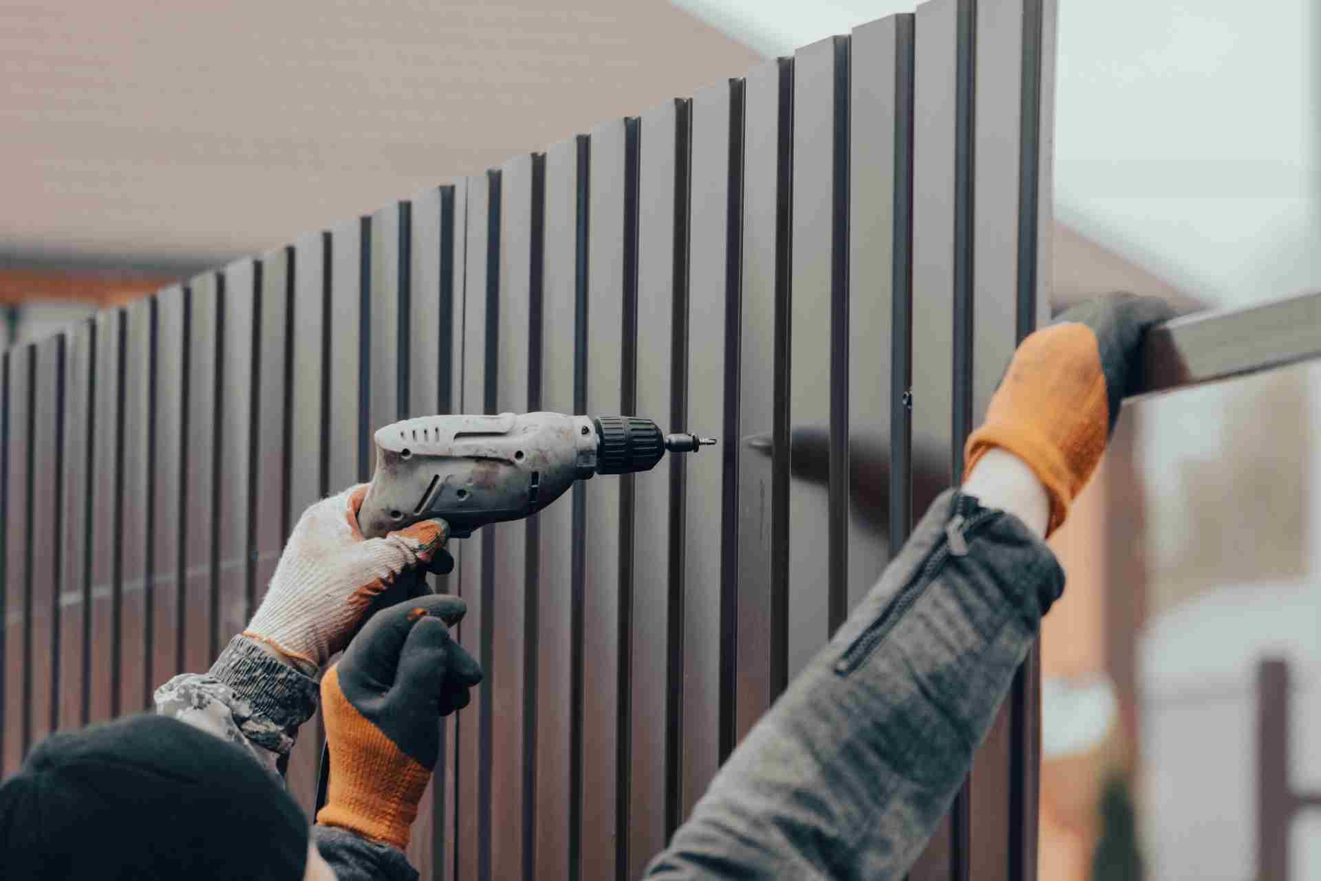 A person wearing work gloves uses a power drill to install brown corrugated metal panels on a fence.