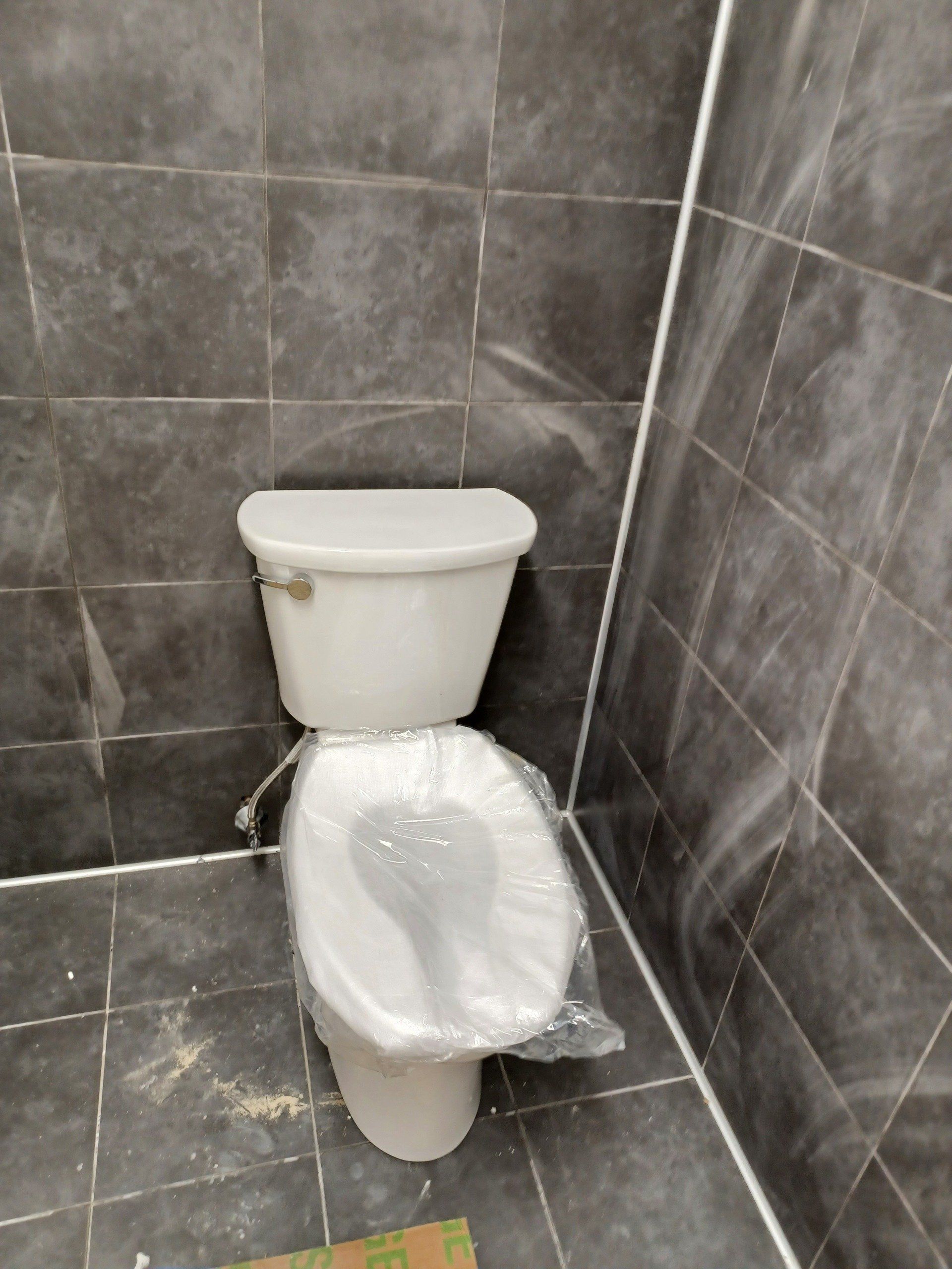 White toilet in a bathroom with dark gray tiled walls and floor, still covered in plastic.
