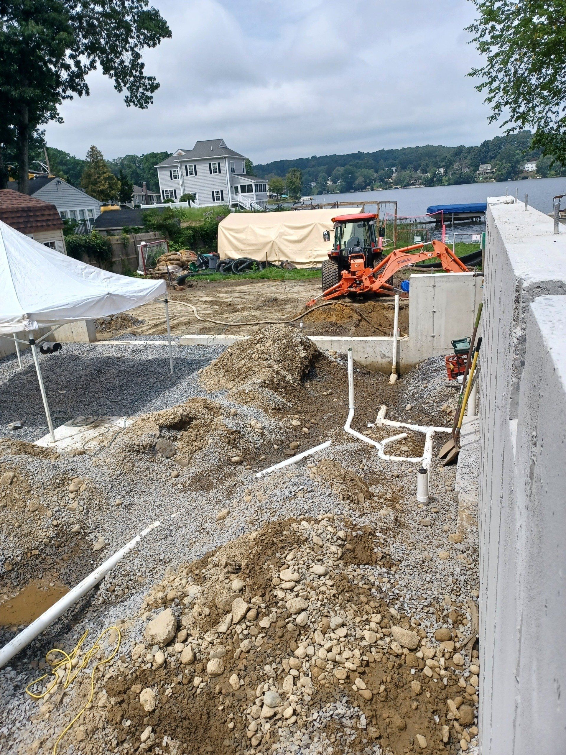 Construction site with backhoe, dirt piles, white foundation, and lake in the background.