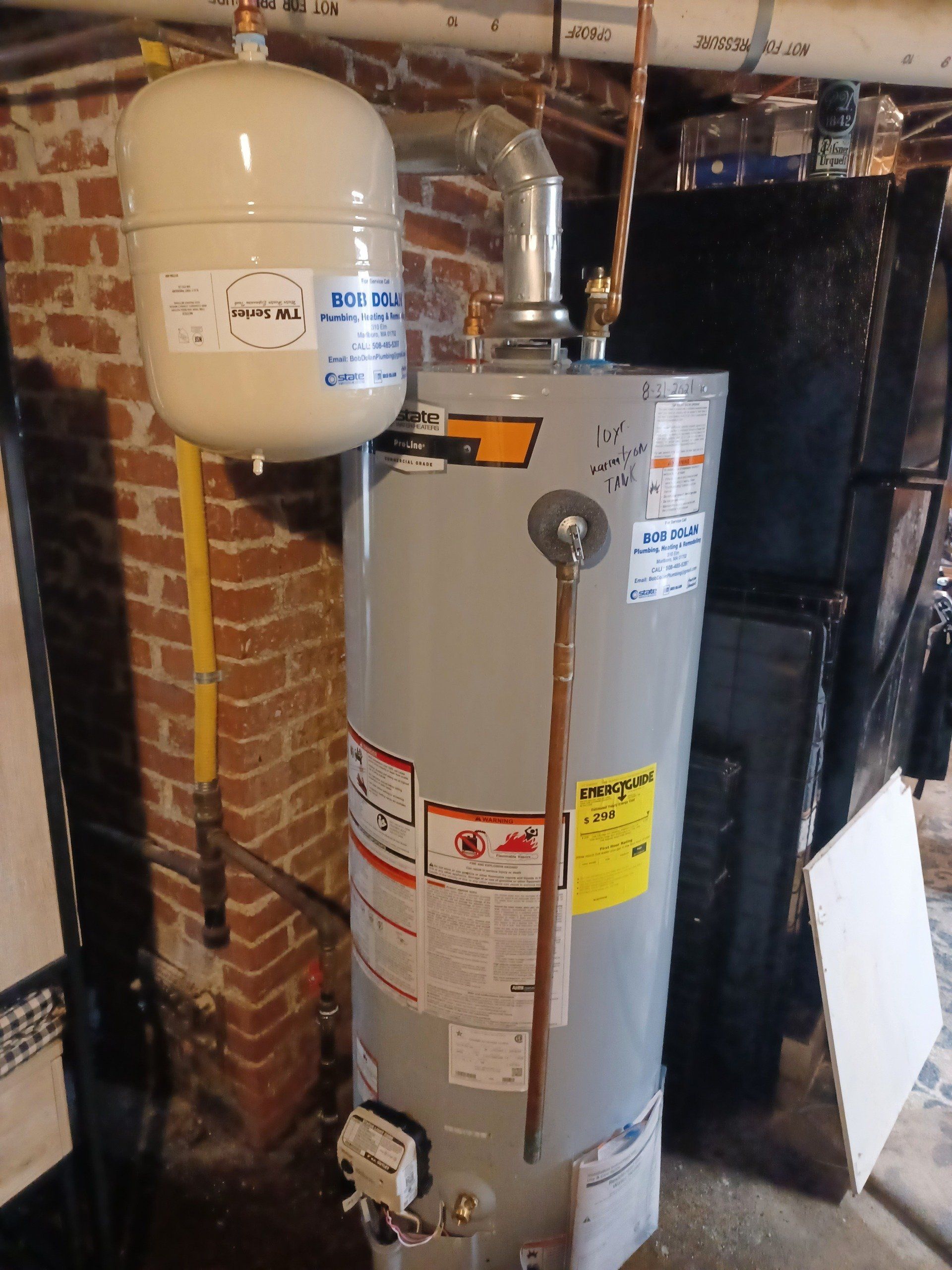 A tall, gray water heater with a white expansion tank and copper pipes against a brick wall.