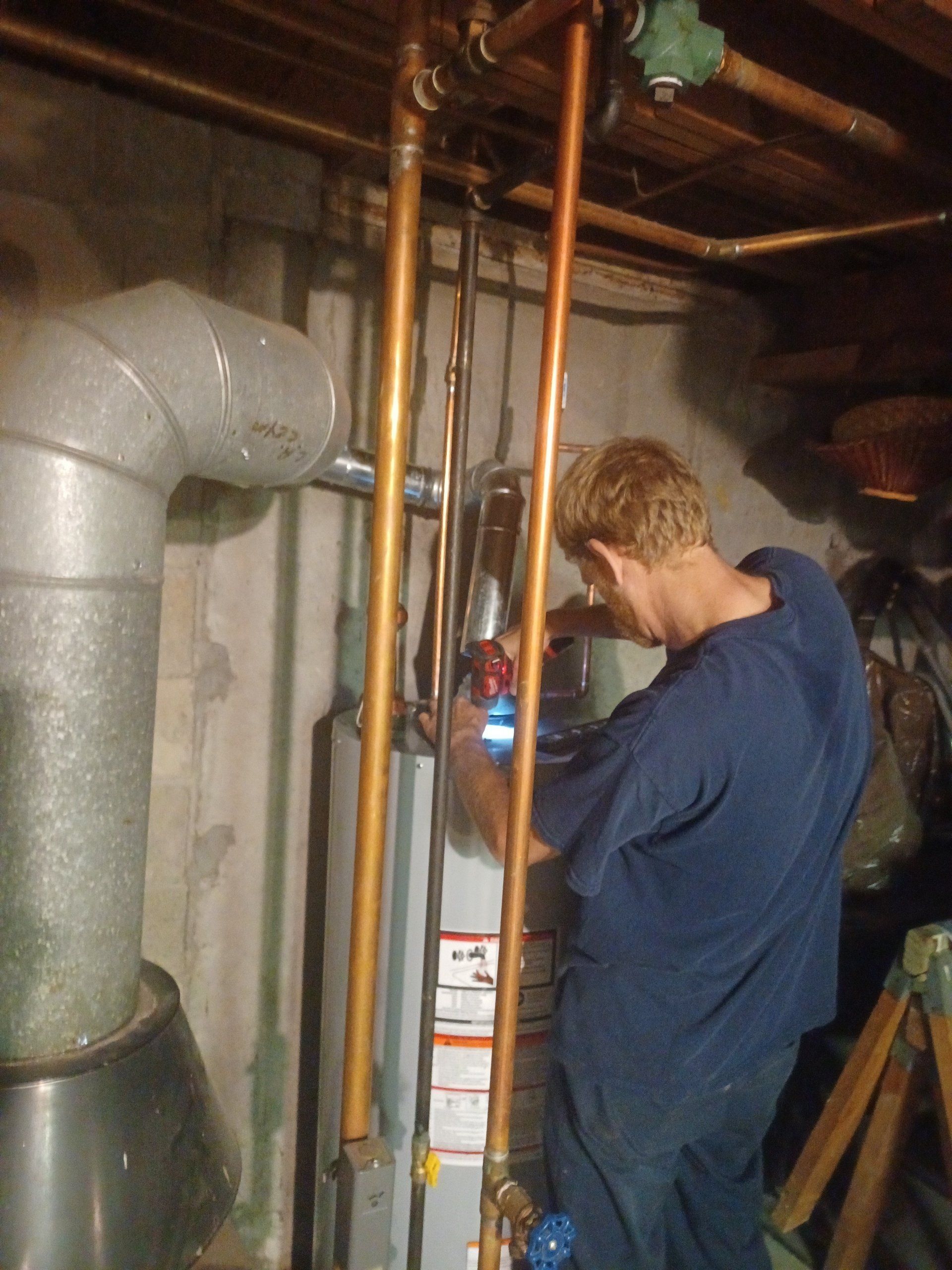 Man in blue shirt working on copper pipes in a basement, near a water heater and furnace.