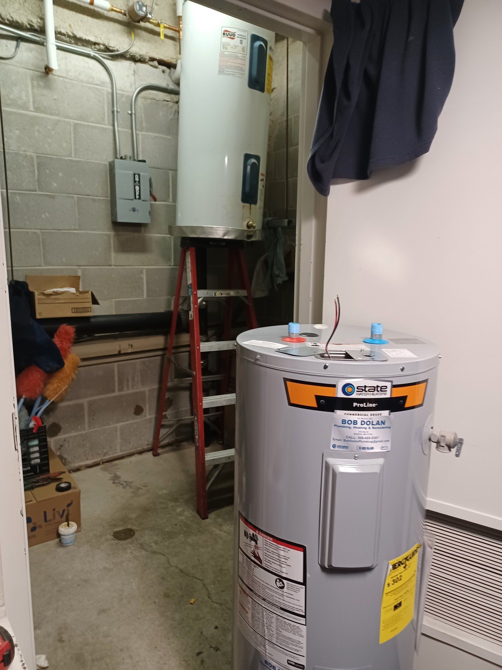 Two water heaters, one new and one on a ladder, in a utility room. Gray cinder block walls.
