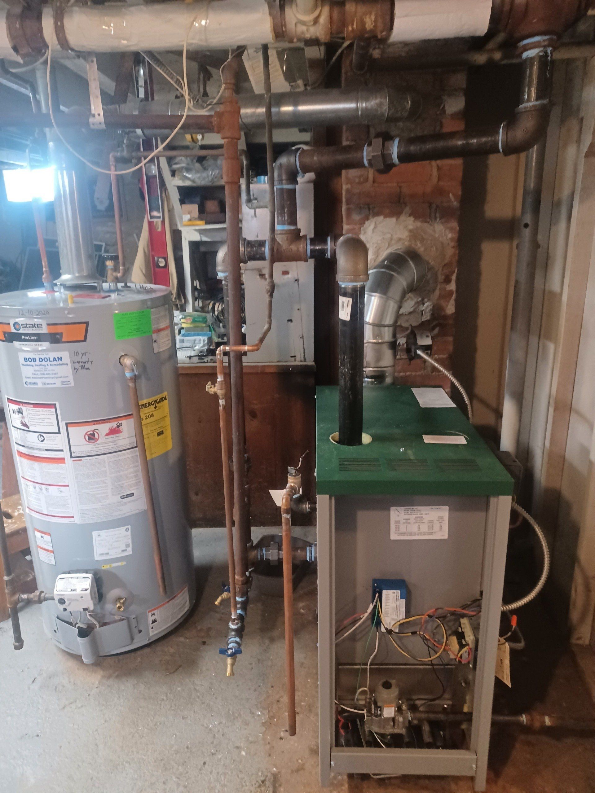 A basement with a hot water heater, boiler, and pipes against a brick wall.