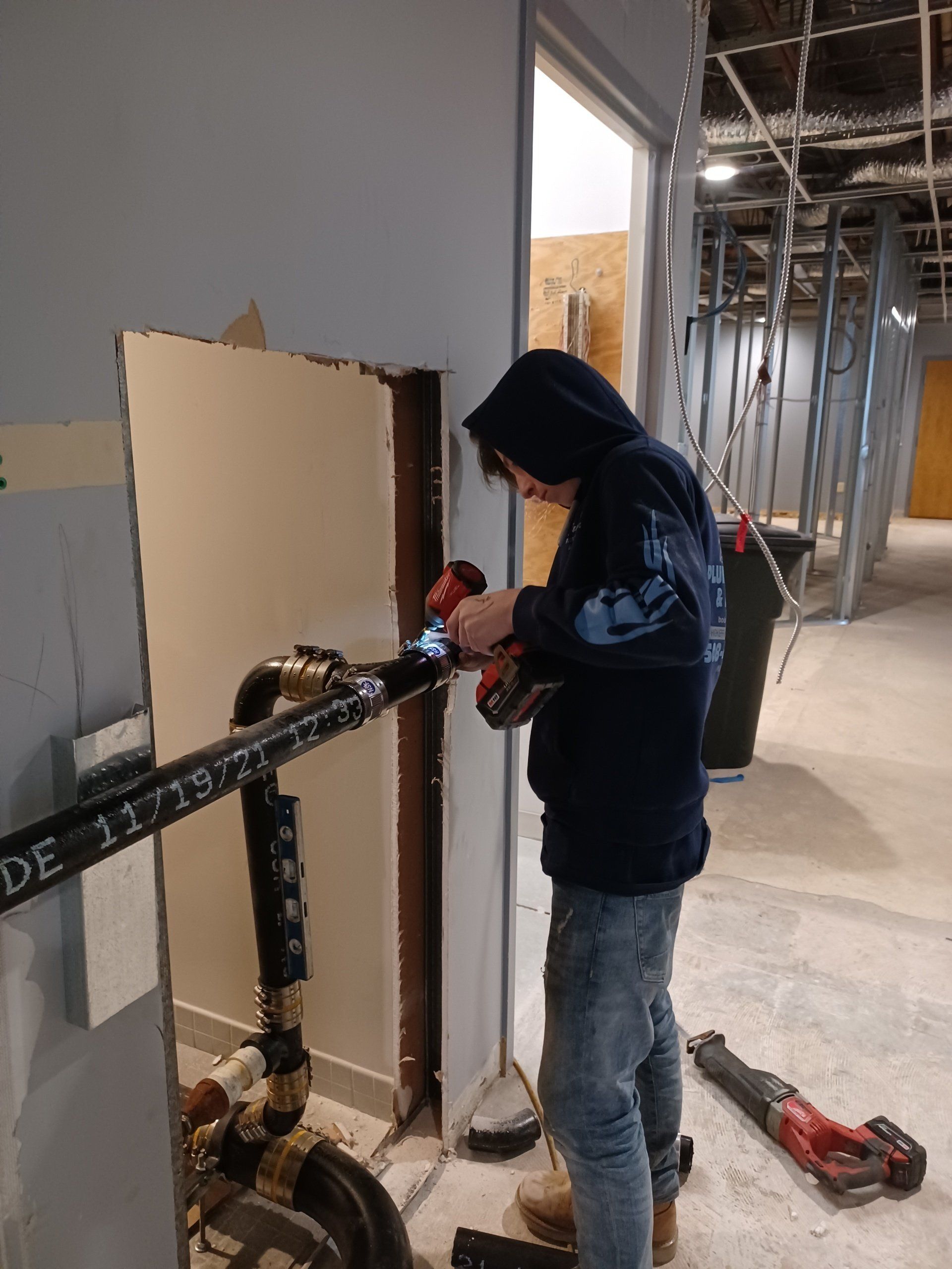 A person in a blue hoodie cutting a black pipe near a doorway in a construction site.