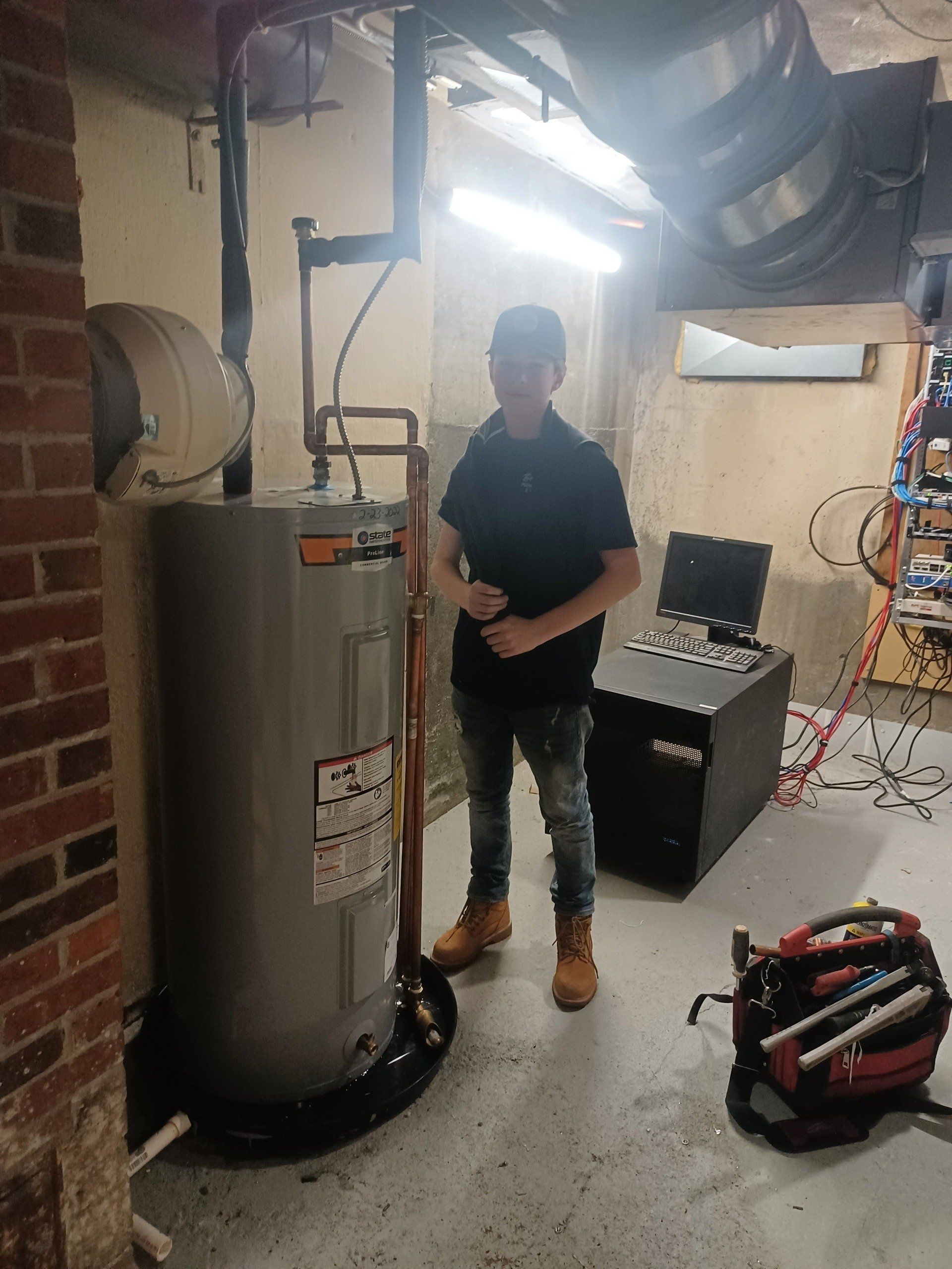 Person in a basement stands near a water heater, looking at the camera. Tools on floor.