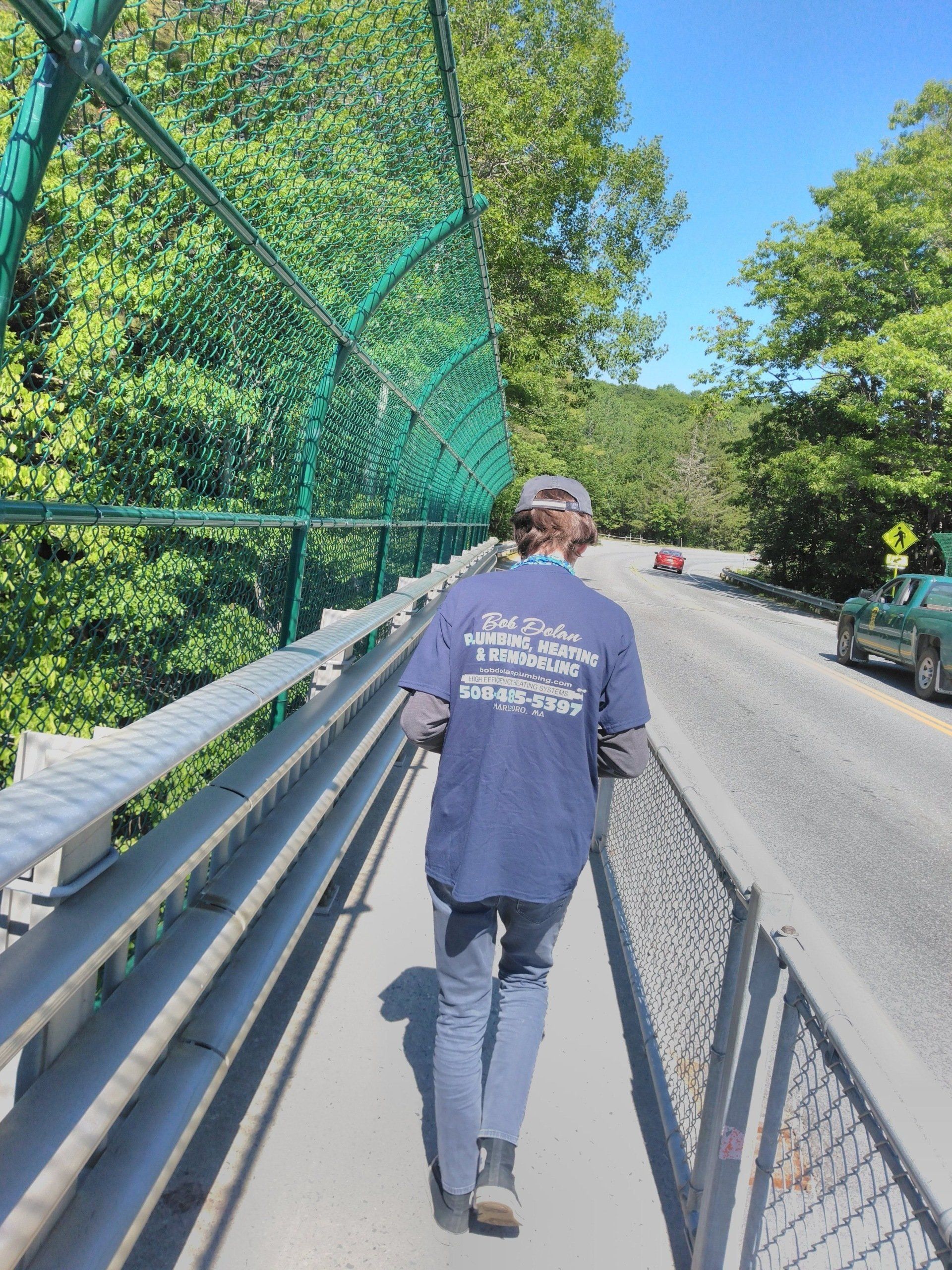 Person in blue shirt walks on bridge with green netting, road, and trees.