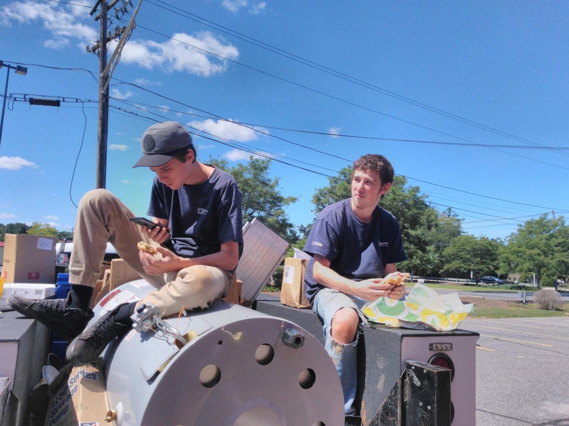 Two young men eating sandwiches, sitting outside on machinery under a blue sky.