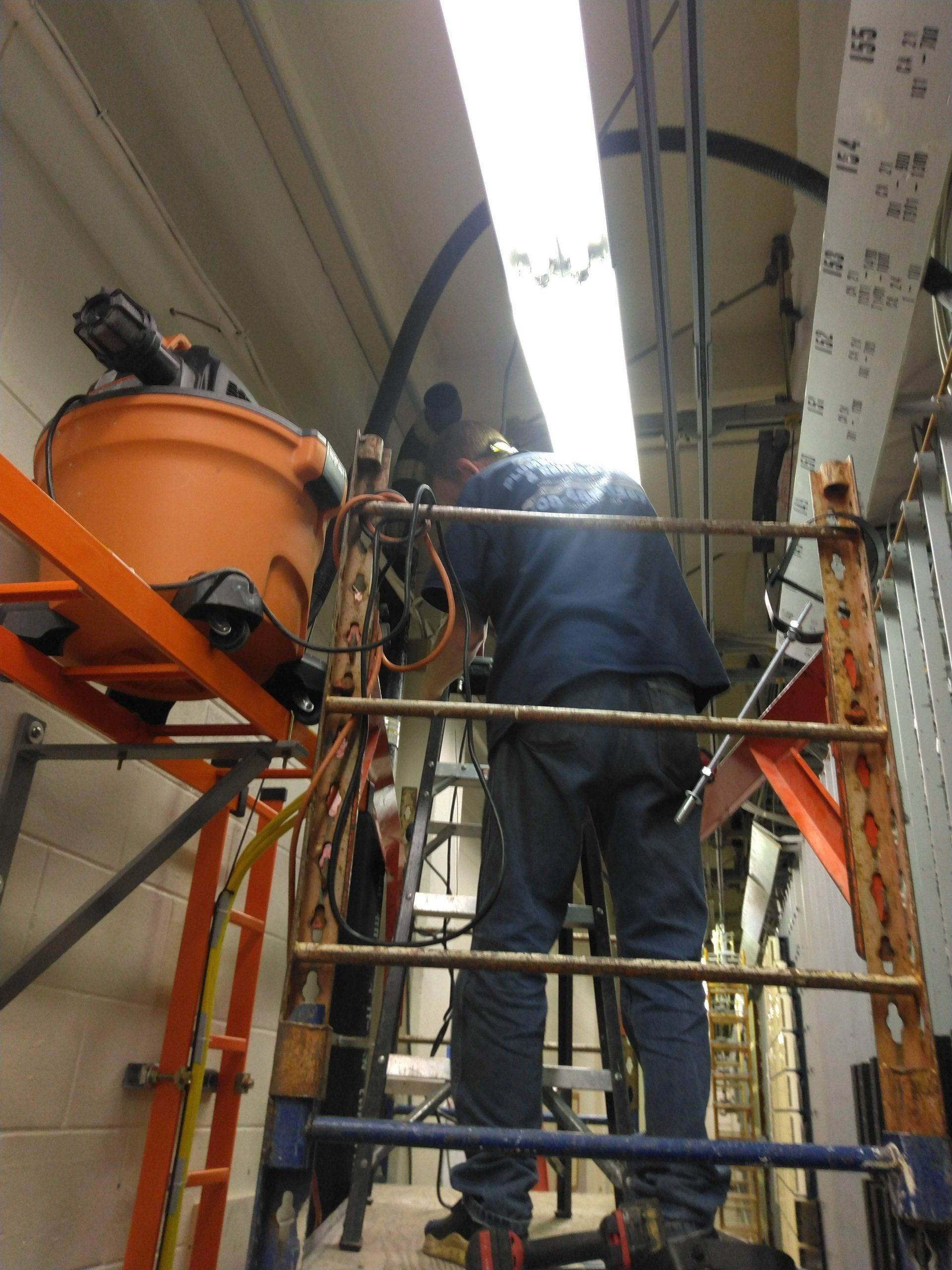 Worker on a tall ladder in a narrow room, wiring cables near overhead lights, with an orange container.