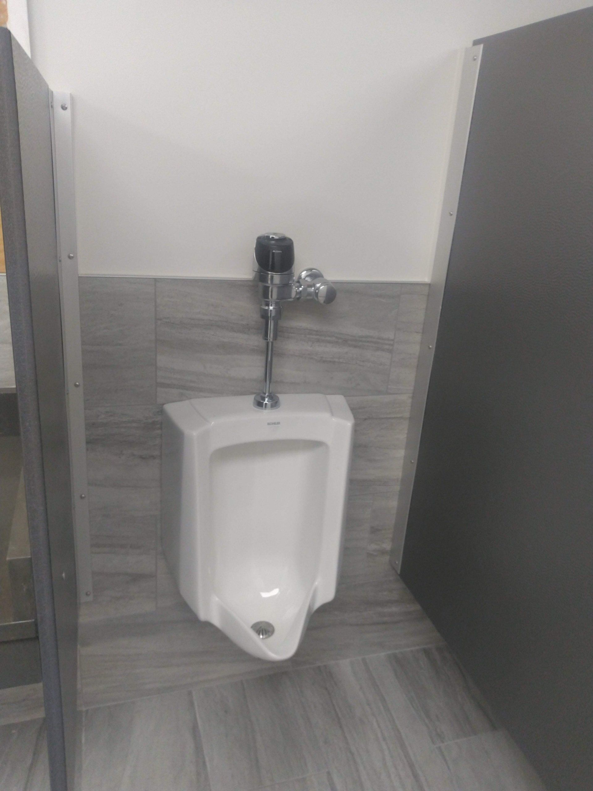 White urinal in a public restroom, gray tile wall and floor, metal flush valve.