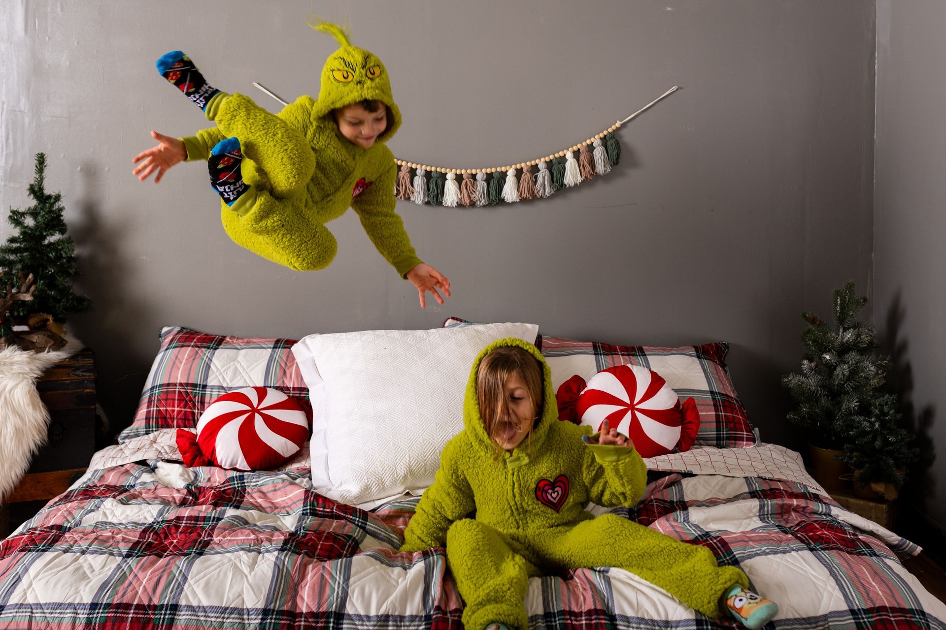 Two children in grinch costumes are jumping in the air while sitting on a bed.