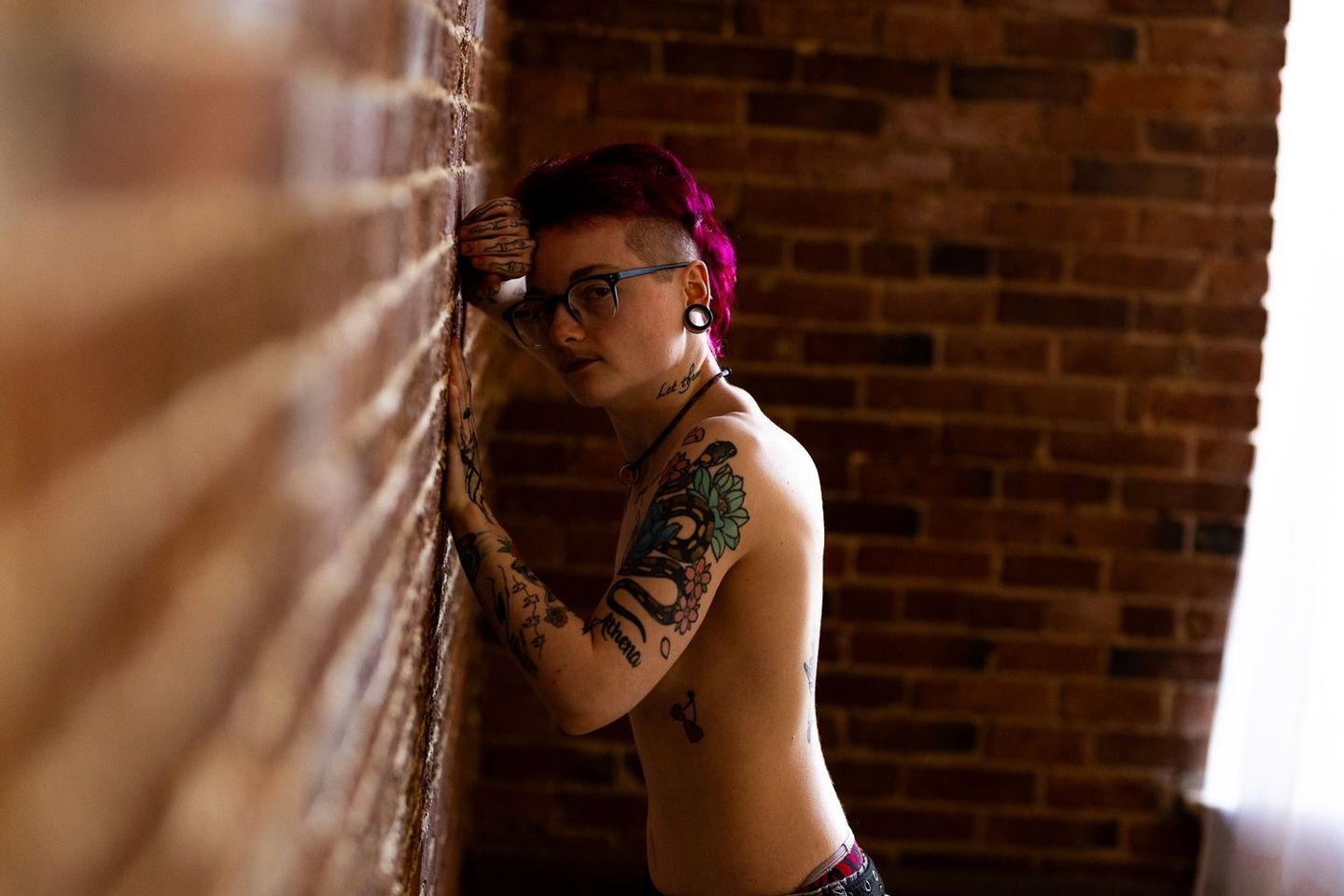 A shirtless trans man with tattoos on his arms leaning against a brick wall for a male boudoir session