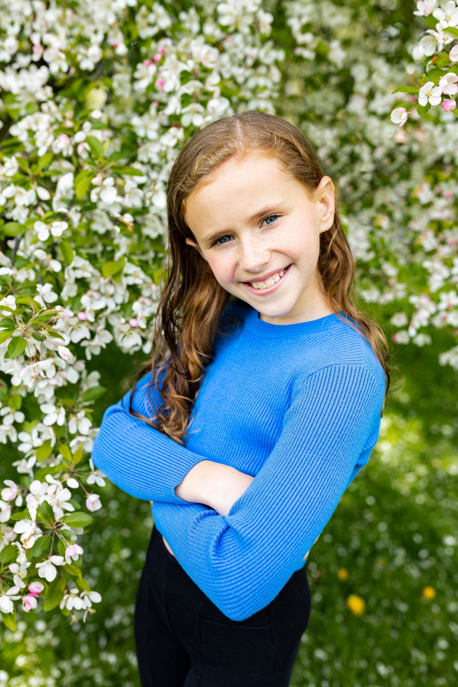 A young girl in a blue sweater is standing in front of a tree with white flowers.