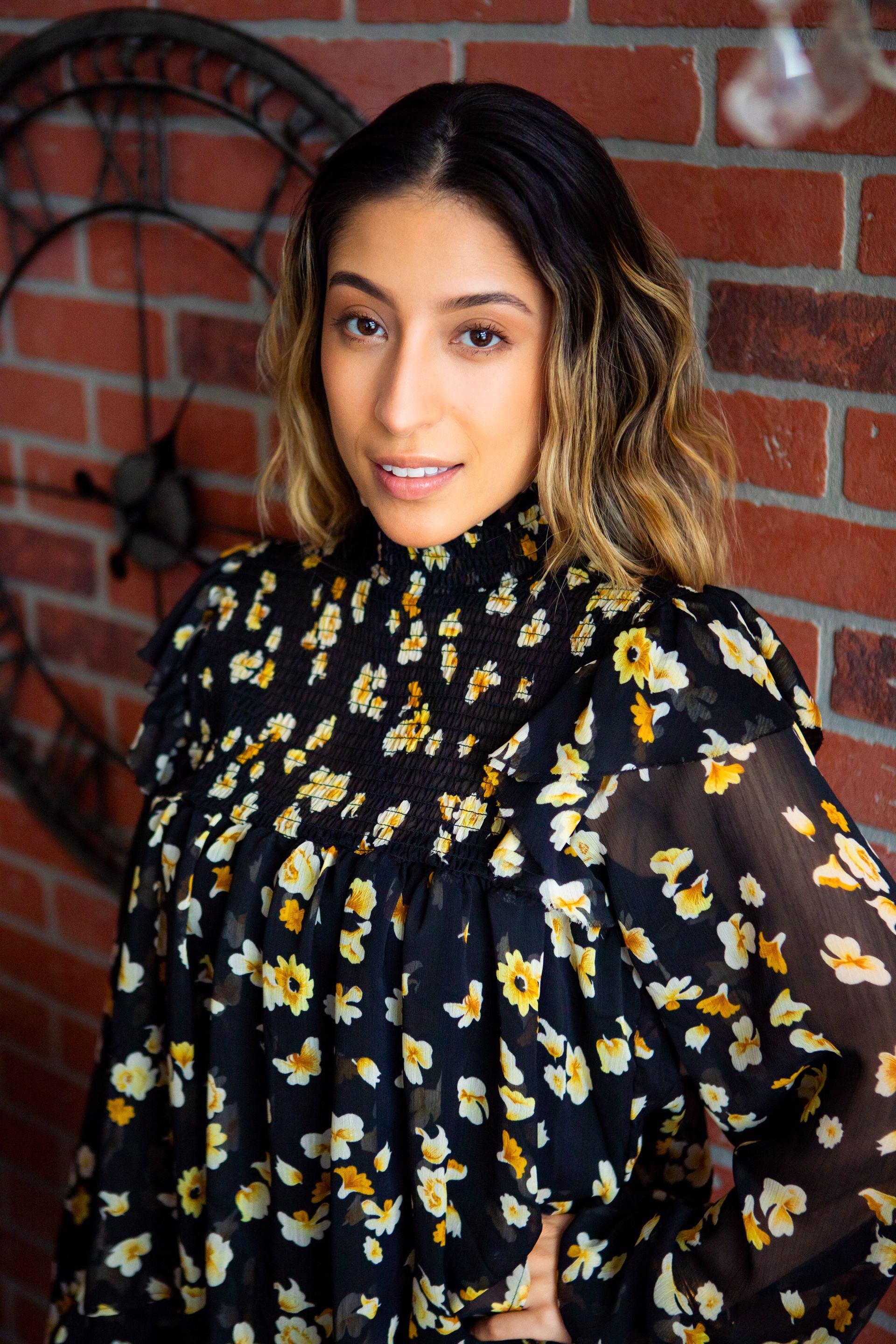 A woman in a floral dress is standing in front of a brick wall.