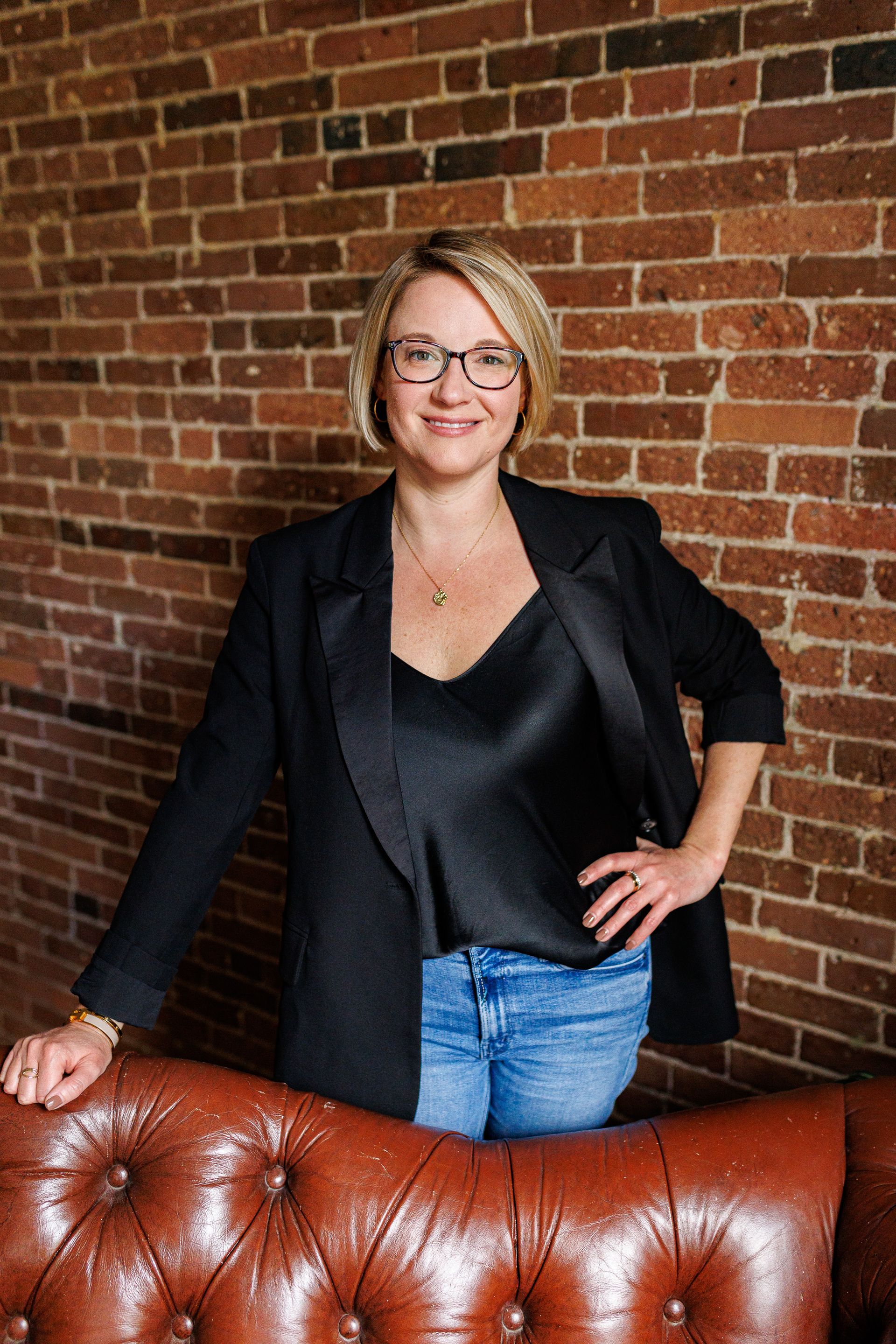 A woman is standing next to a leather couch in front of a brick wall.