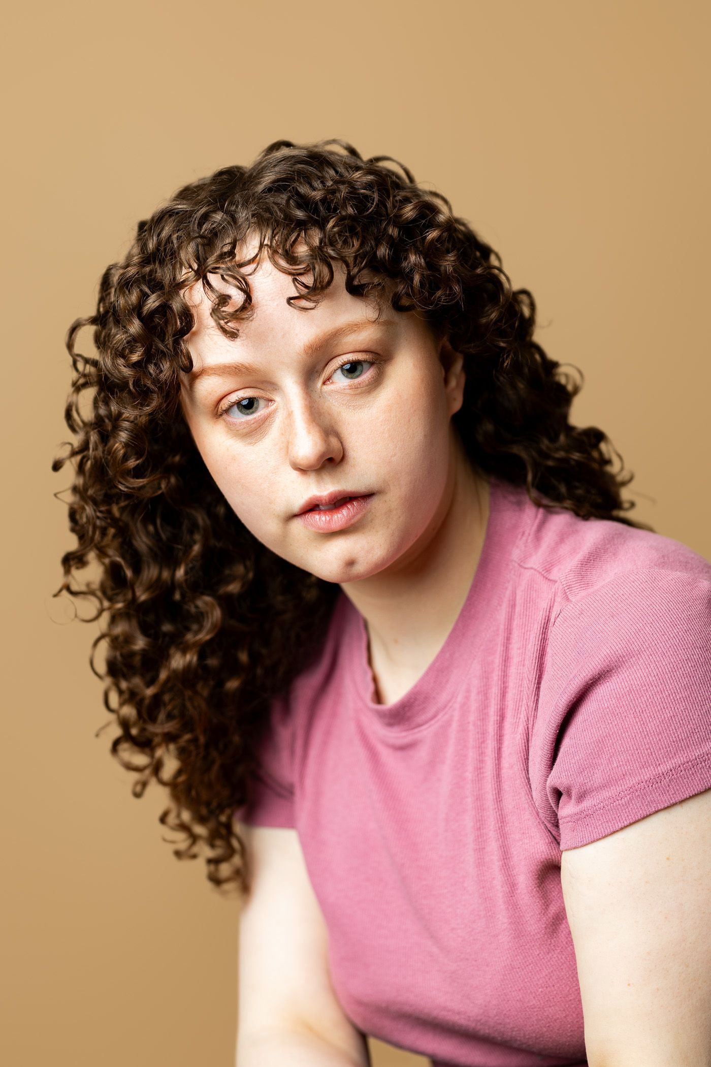 A woman with curly hair is wearing a pink shirt and looking at the camera.