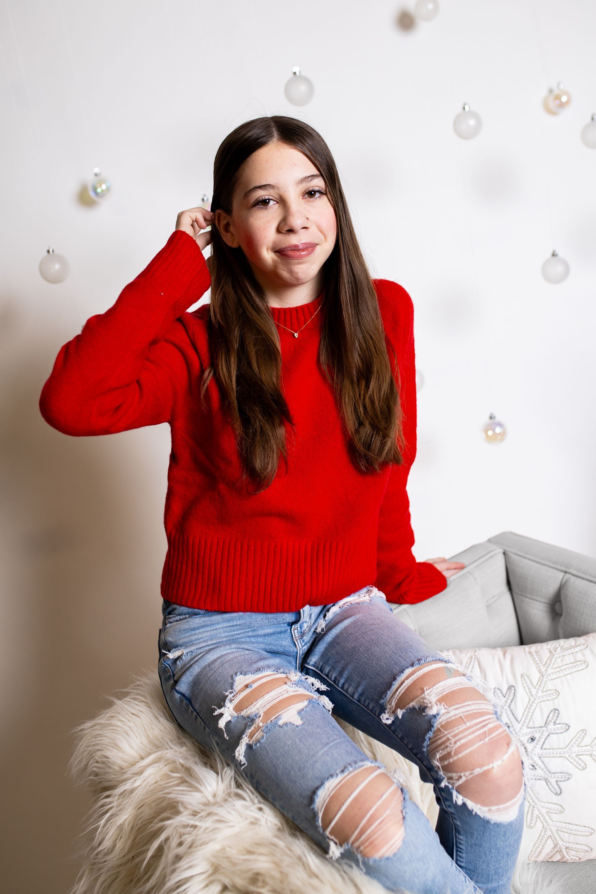A young girl in a red sweater and ripped jeans is sitting on a couch.