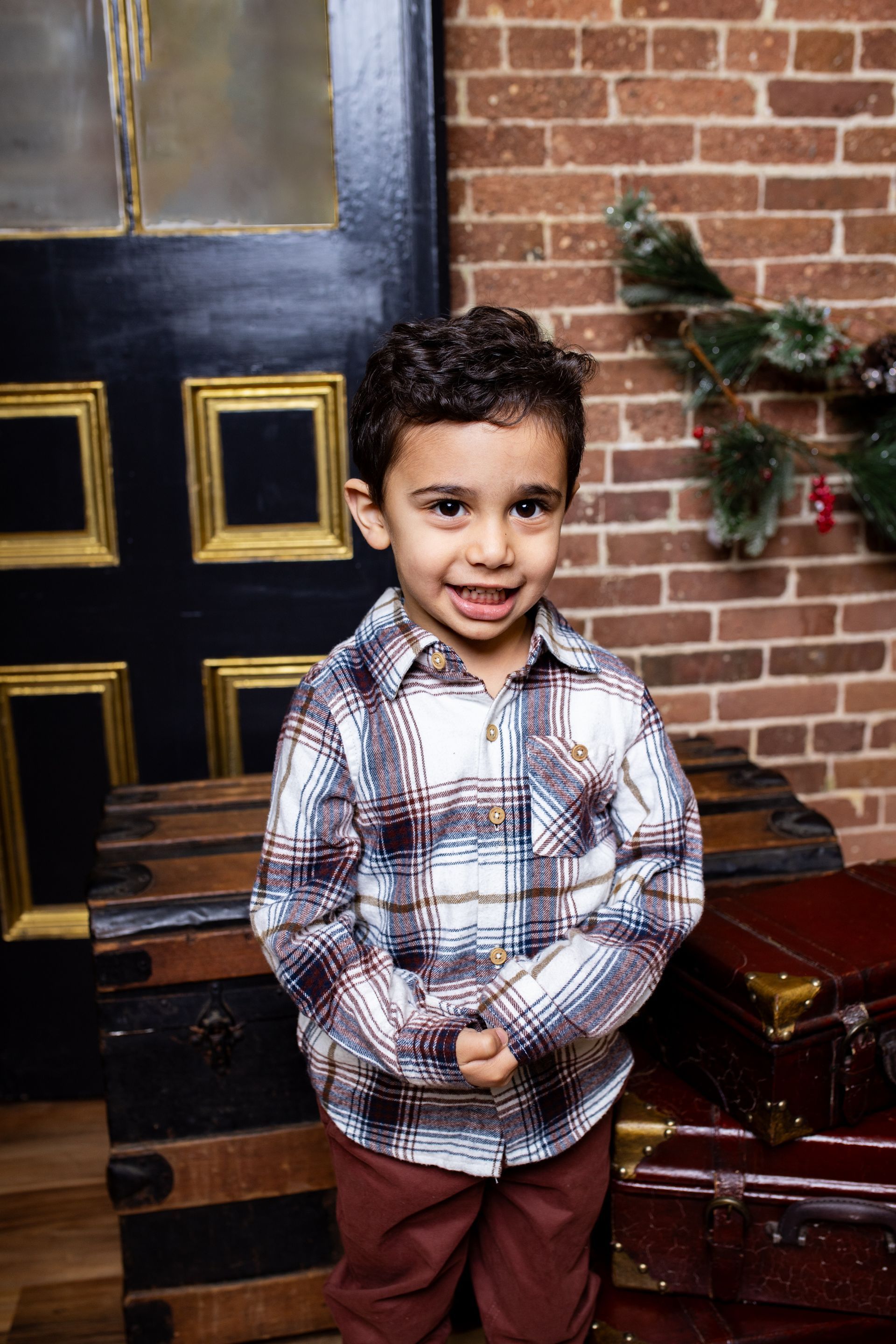 A little boy in a plaid shirt is standing in front of a brick wall.