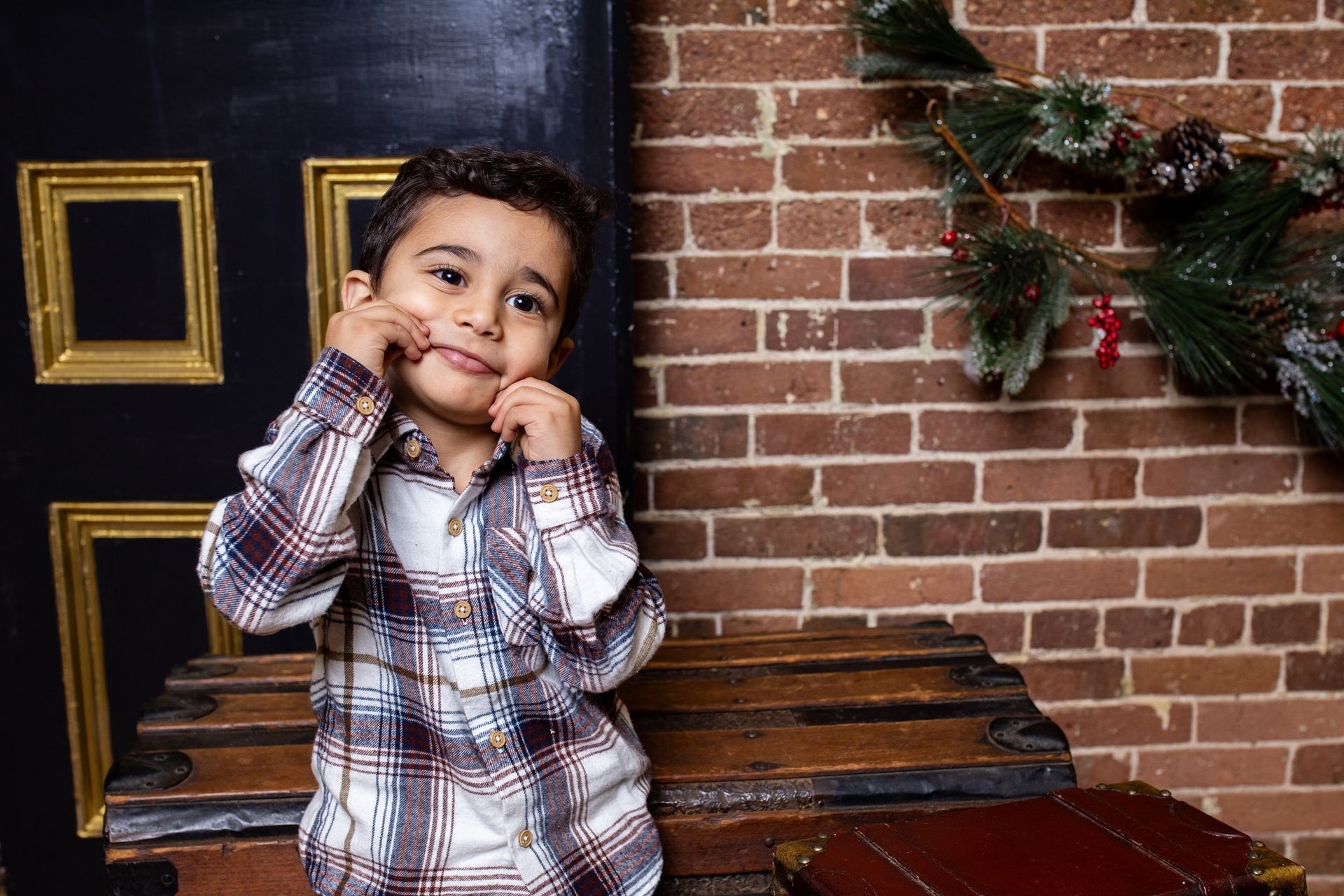 A little boy in a plaid shirt is standing in front of a brick wall.