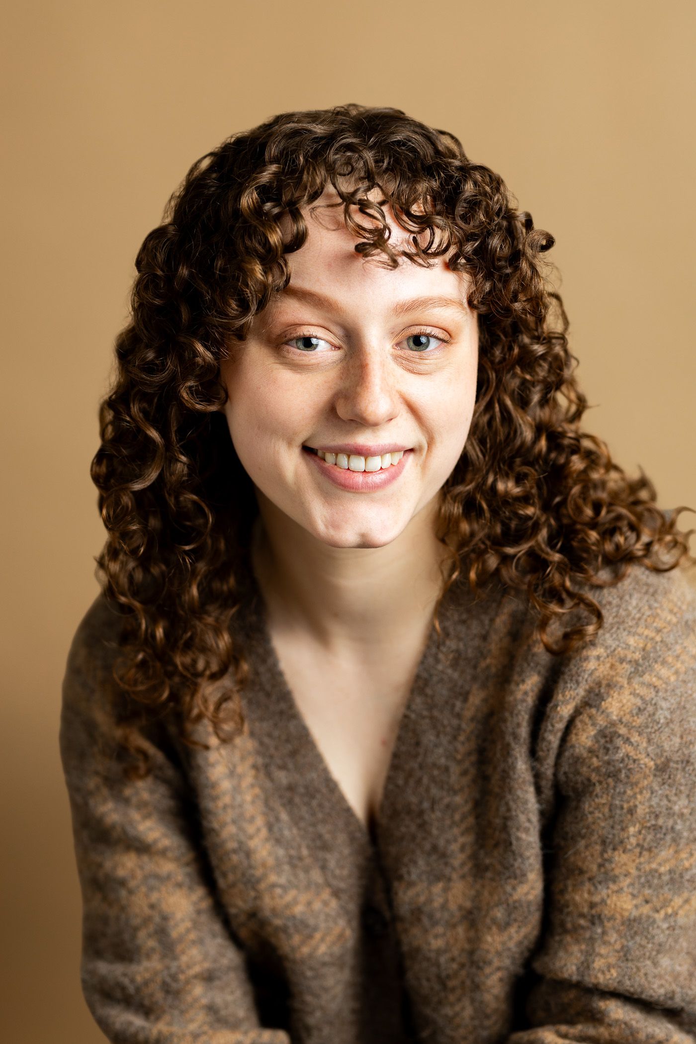 A young woman with curly hair is smiling and wearing a brown sweater.