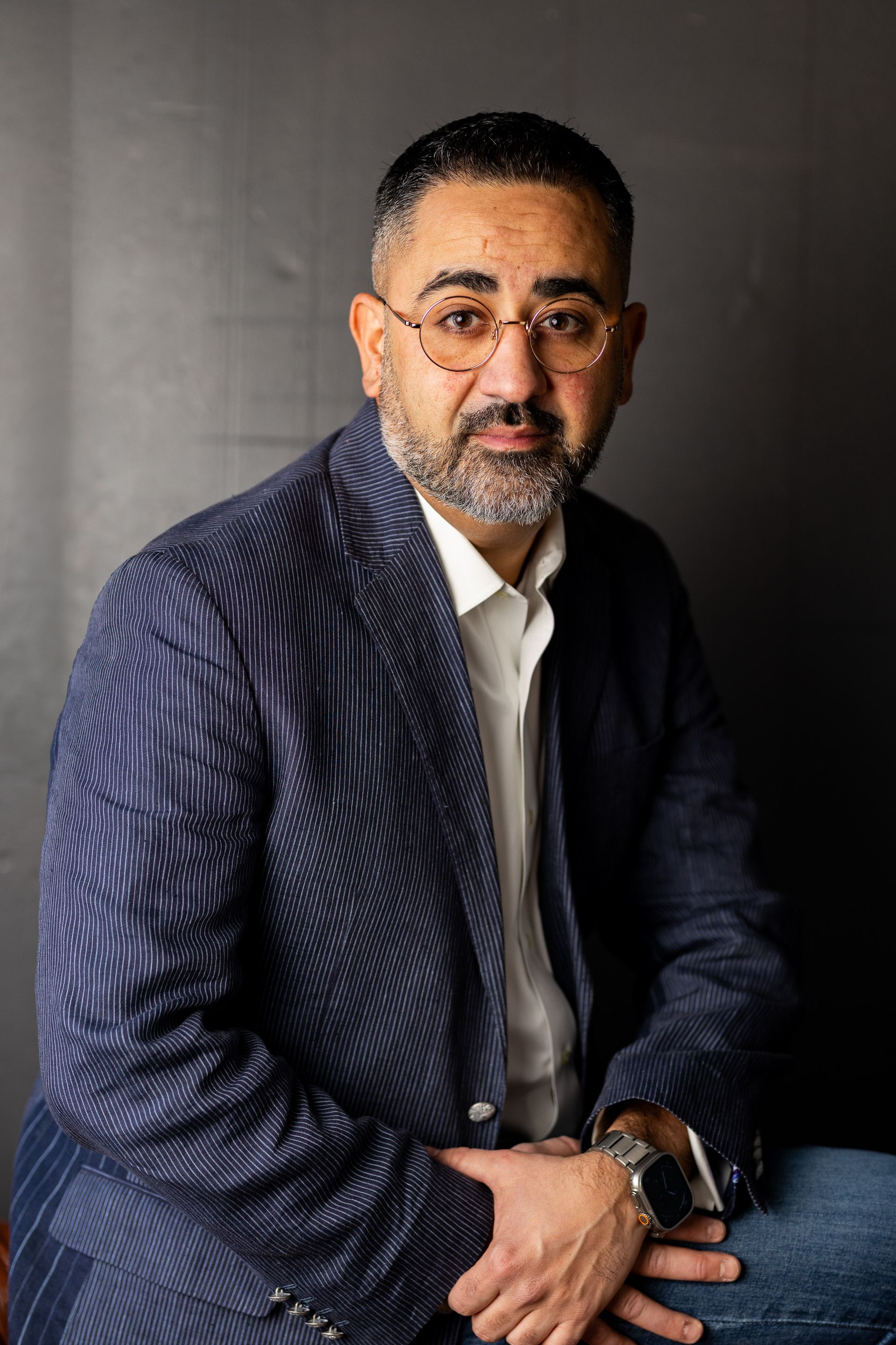 A man with a beard wearing glasses and a blue jacket is sitting down.