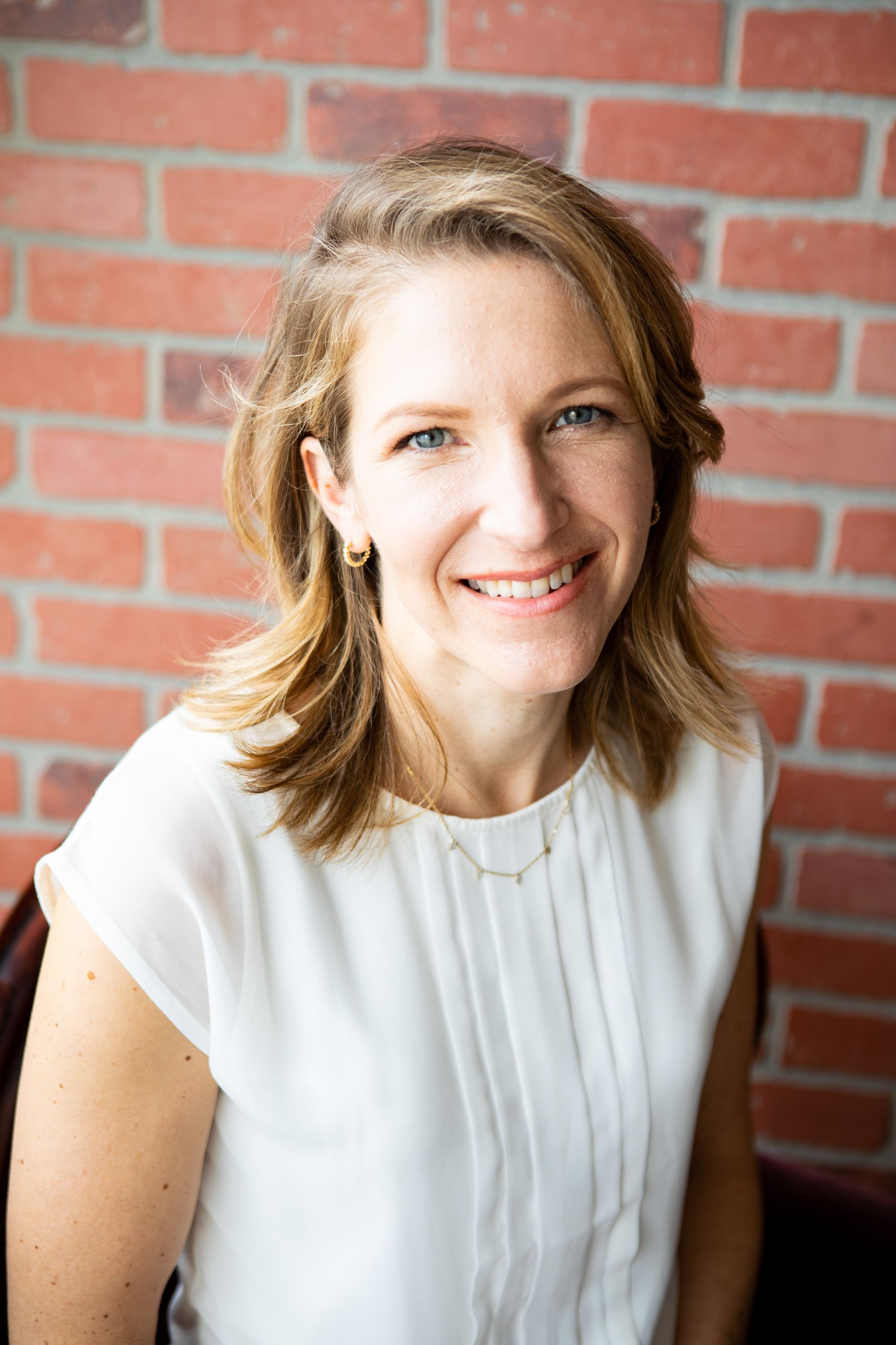 A woman in a white shirt is smiling in front of a brick wall.