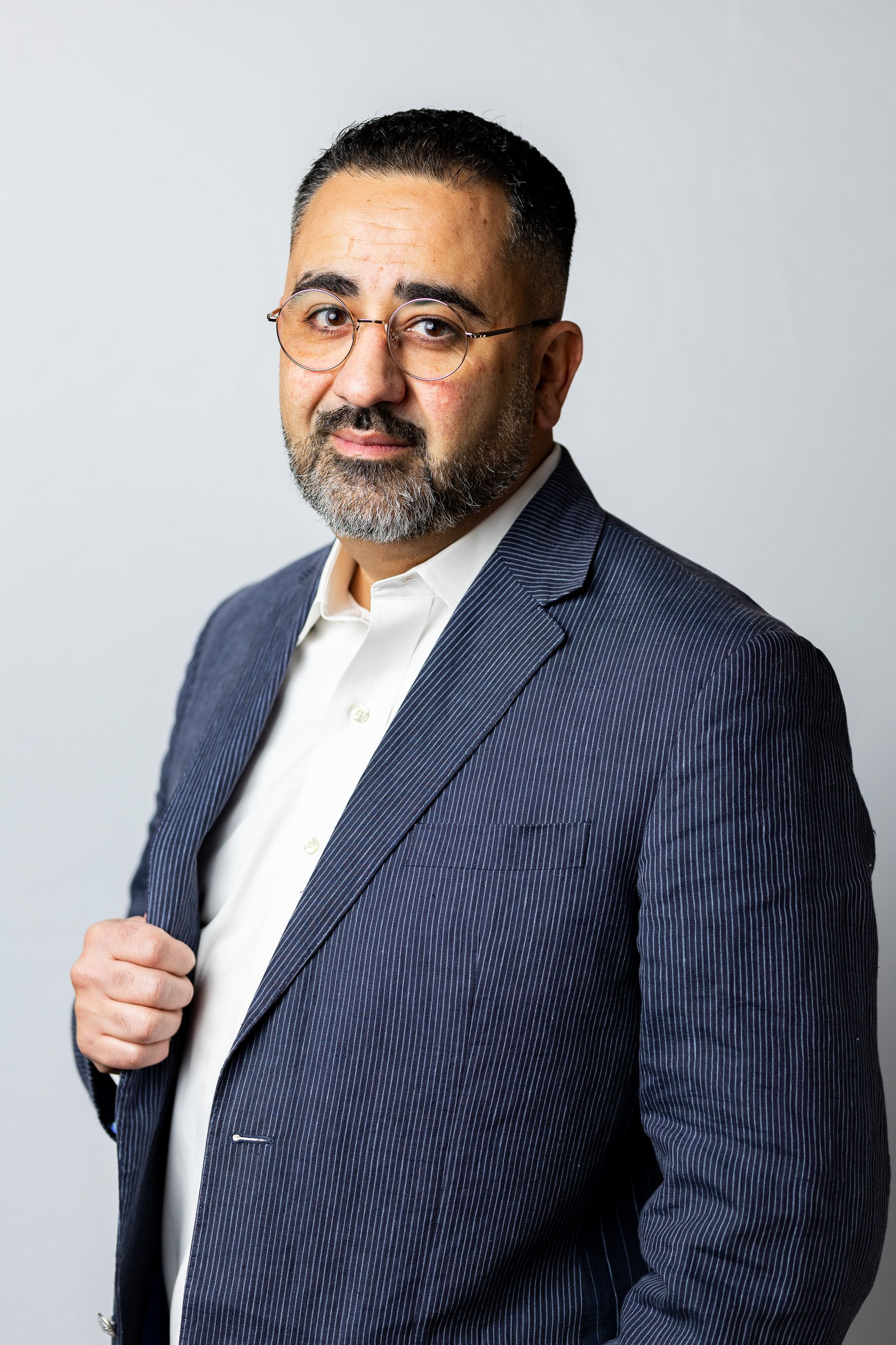 A man in a suit and glasses is standing in front of a white wall.