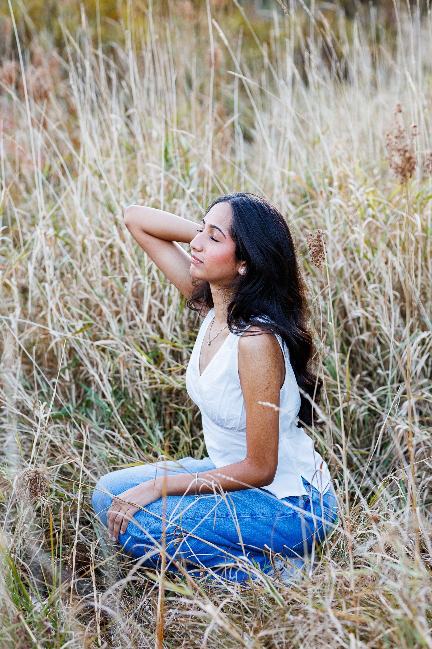 Photography MA -A woman is sitting in a field of tall grass with her eyes closed.